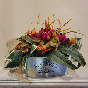 Orange and red toned flowers with long green leaves in a "Flowers & Garden" silver dish