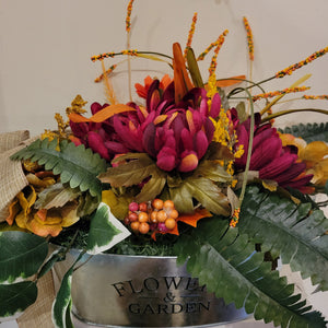 A close up of the orange and red toned flowers with long green leaves in a "Flowers & Garden" silver dish