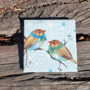 A 4x4 coaster with a painting of two brown birds on a snowy branch wrapped in blue and green scarves.