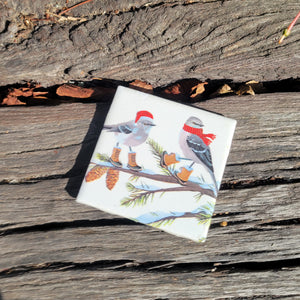 A 4x4 coaster with a painting of two grey birds with tan boots on a snowy branch. The left bird is wearing a red santa hat and the right bird is wearing a red scarf.