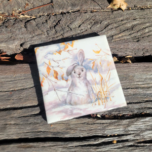 A 4x4 coaster with a painting of a white bunny in the snow with one ear down.