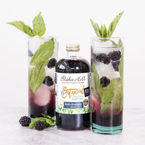 The Basil & Blueberry shrub in between two glasses filled with liquid and basil