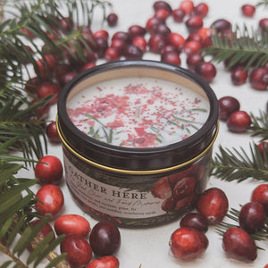Ash & Rose candle surrounded by cranberries and pine trees. The candle has cranberry powder and pines sticking out of the top.