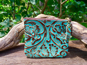Turquoise coin purse with zip top leaning againsta  piece of driftwood with greenery backdrop.