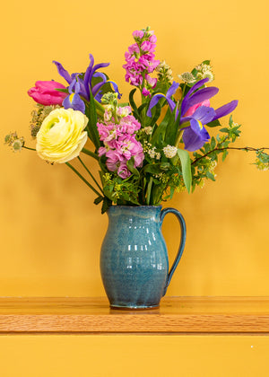 Blue stoneware pitcher in blue glaze with graceful handle holding assorted fresh flowers sitting on a shelf with yellow background.